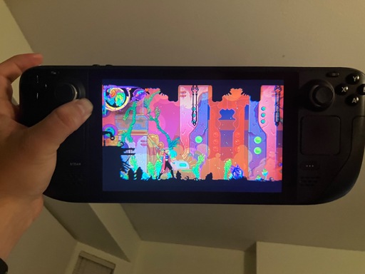 A person is holding up a Steam Deck, showing a colorful 2D game on its screen. The game features a surreal, neon-hued environment with pink, orange, and purple tones, green vines, glowing orbs, and silhouetted characters along the bottom edge. The vivid psychedelic art style fills the display, contrasting with the dark handheld console casing.