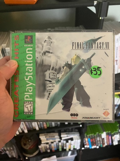 A hand holds a PlayStation 1 “Greatest Hits” edition copy of Final Fantasy VII. The cover shows Cloud Strife with his massive Buster Sword slung over his shoulder, facing the Shinra building in Midgar. The green “Greatest Hits” banner runs along the left side of the jewel case, with the PlayStation logo and “Teen” ESRB rating visible. A bright green price sticker reads “$35”. The case is slightly scuffed with a crease running across the lower part of the insert. Shelves of other video games are visible blurred in the background.