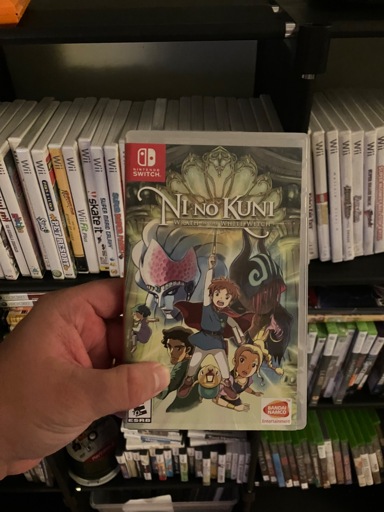 A person is holding a Nintendo Switch game case for Ni no Kuni: Wrath of the White Witch. The cover features colorful anime-style artwork with the main character Oliver in the center, raising a wand while surrounded by companions and fantastical creatures. The Bandai Namco Entertainment logo appears in the bottom right corner. Behind the case, shelves of neatly arranged Wii game cases are visible, along with more games stored below.