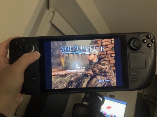 A person holds a Steam Deck handheld gaming PC showing the title screen of Sniper Elite. The display features a soldier in camouflage crouched beside a brick wall, holding a sniper rifle, with the game’s title in bold blue letters above him. The text “Start Game” appears in the bottom right of the screen. In the background, a desk with a monitor and chair is visible, slightly out of focus.