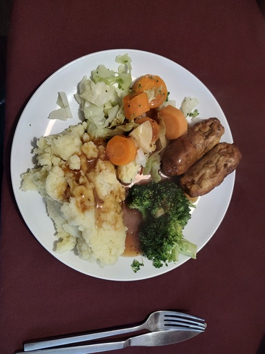 A plateful of vegan bangers, vegetables - carrots, cabbage, broccoli - with a big dollop of mashed potatoes, smothered in vegan gravy.