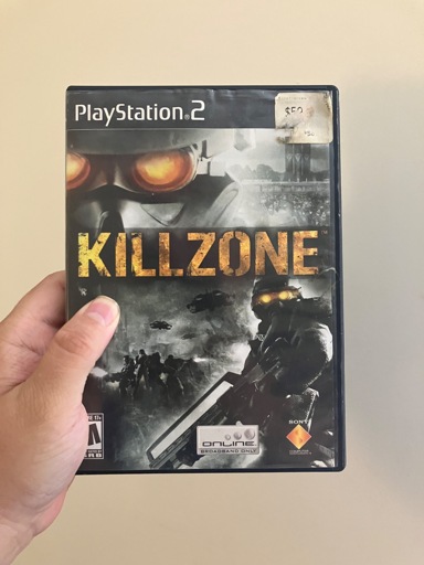 A person holding a PlayStation 2 copy of Killzone. The cover shows a Helghast soldier with glowing orange eyes in the background, and another armed Helghast in the foreground, standing before a smoky battlefield with helicopters and troops. The title “KILLZONE” is printed in bold, distressed yellow letters across the middle. A faded price sticker in the top right corner lists it as $5.00.