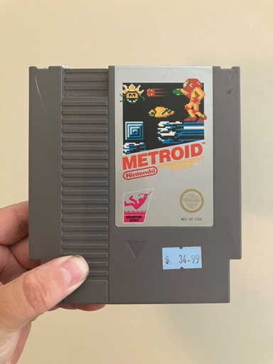 A person is holding a gray NES cartridge of Metroid. The label shows pixel art of Samus firing her arm cannon, with enemies and a sci-fi background. The cartridge features the Nintendo “Adventure Series” logo and gold seal of quality. A price sticker on the front reads $34.99.